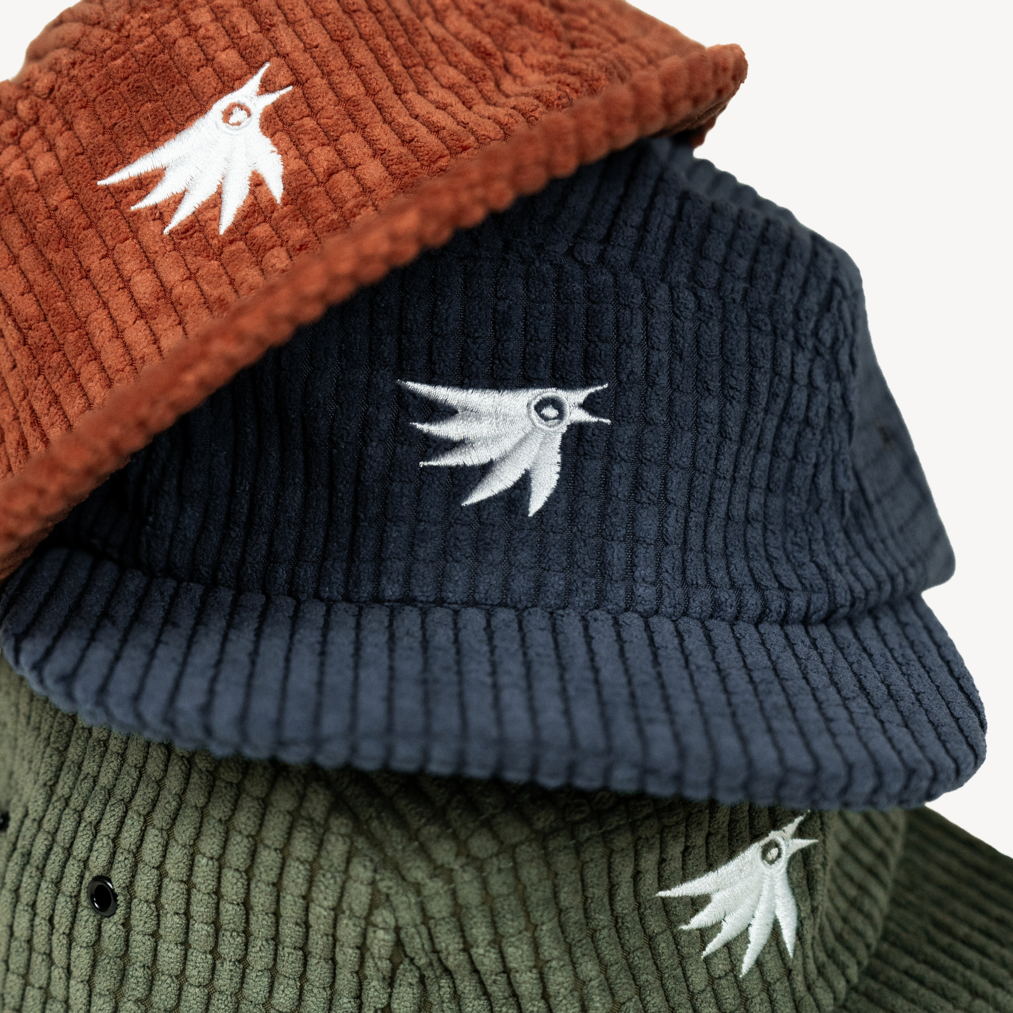 Three ribbed corduroy flat-brim hats stacked together, showing Canyon Amber, Midnight Blue, and Evergreen Pine colors, each with a white embroidered bird logo on the front.